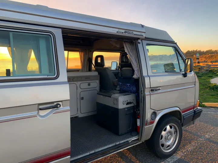 An RV rental, a vintage Westfalia camper van, sits by the coast at sunset, revealing its open door interior and ocean view.. Volkswagen T3 Westfalia 1991