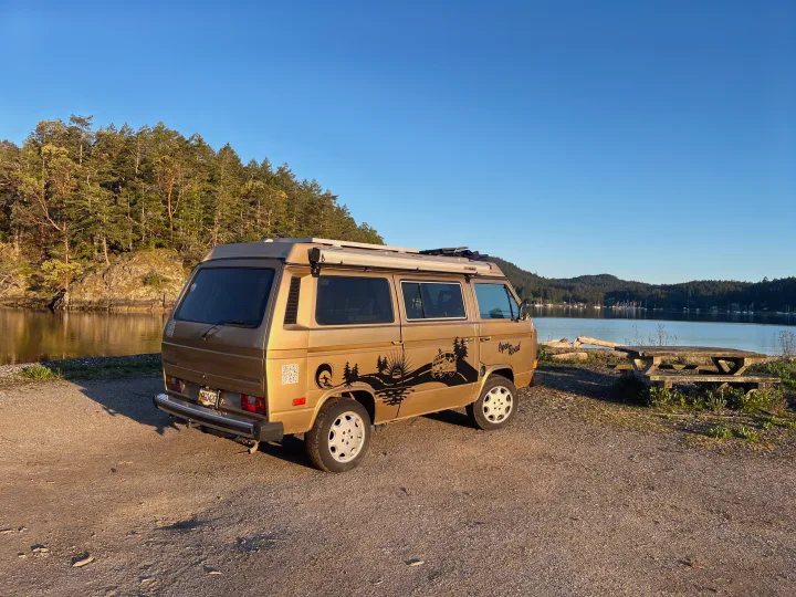 Golden VW campervan by a tranquil bay with forested hills & picnic table. Ideal rv rental for nature adventures.. Volkswagen T3 Westfalia 1985