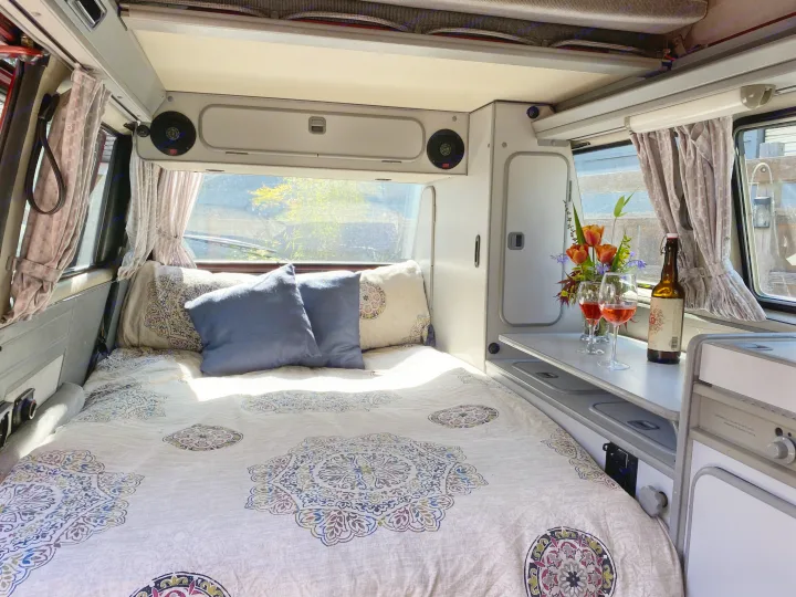 Inviting rv rental interior with a comfortable bed, curtains, and a table featuring drinks and flowers. Ideal for road trips.. Volkswagen Westfalia 1986