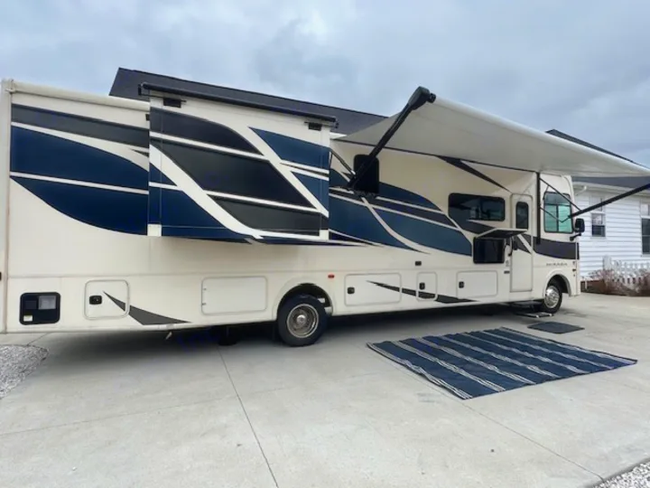 This RV rental shows a cream motorhome with a slide-out and awning extended for outdoor living, parked on concrete with a striped rug.. Forest River Coachmen Mirada 2023