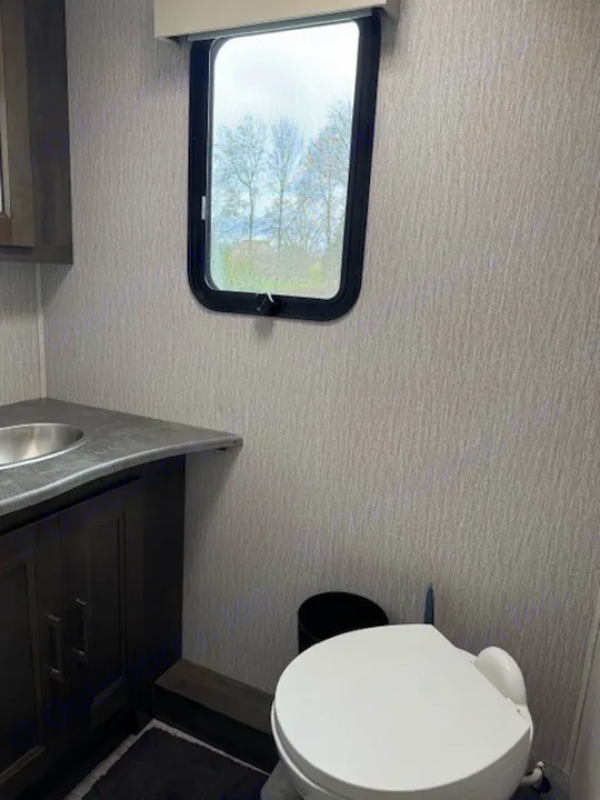 Compact RV rental bathroom showing a white toilet, small sink in a dark vanity, and a window looking out at trees.. Forest River Coachmen Mirada 2023