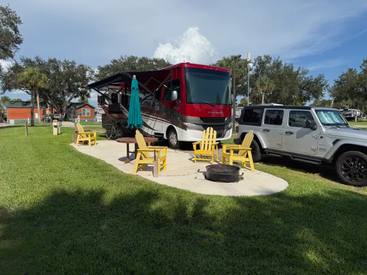 RV rental setup: A red motorhome and silver Jeep parked at a site with yellow Adirondack chairs and a fire pit.. Tiffin Allegro 2020