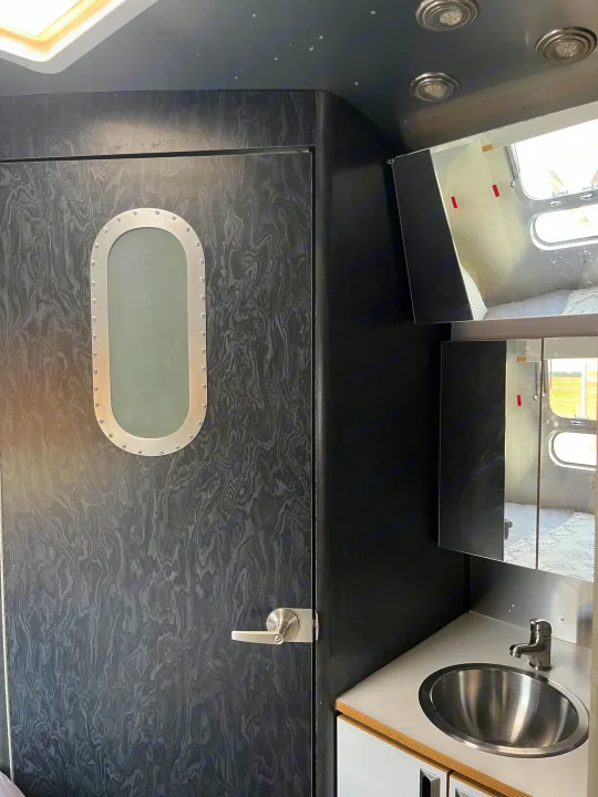 Inside a dark, modern RV rental, showing a porthole door, vanity with a stainless steel sink, and mirrored cabinet.. Airstream Bambi International 2006