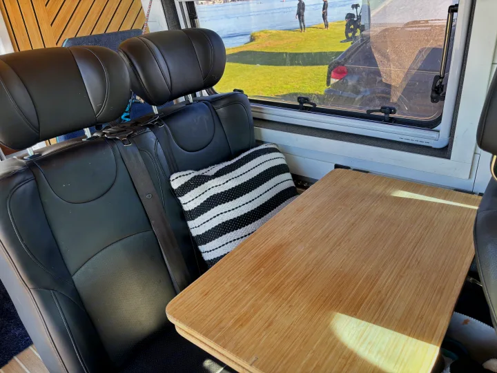 RV rental interior with black seats, a bamboo table, and a window view of wetsuit-clad people by the water.. Mercedes Sprinter 2022