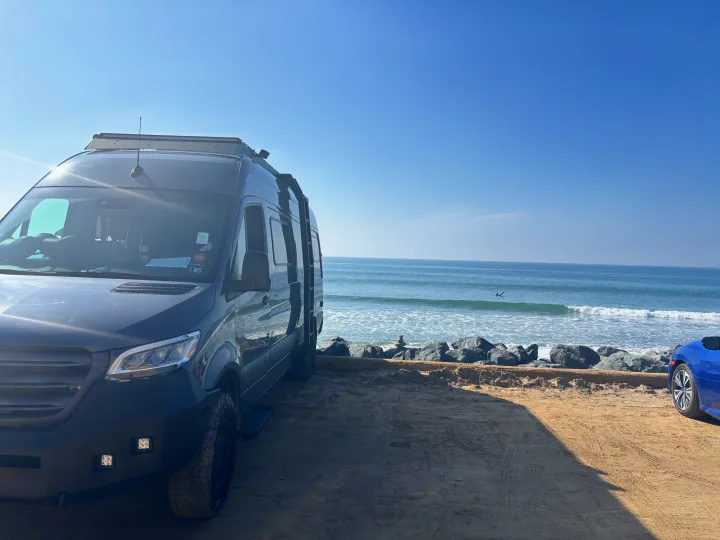 Coastal rv rental: Dark camper van parked on a sandy beach. Ocean waves, clear blue sky, and a distant surfer create a serene scene.. Mercedes Sprinter 2022