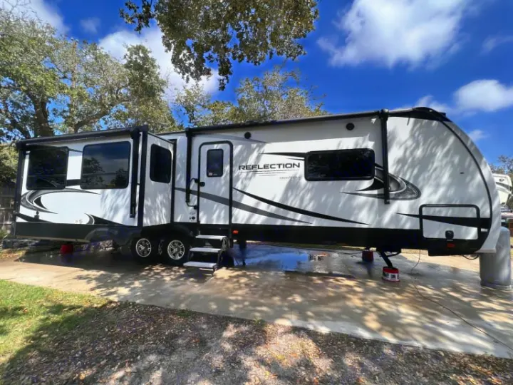A white fifth-wheel RV rental is parked on a concrete slab, steps extended, under trees on a bright day.. Grand Design Reflection 2020