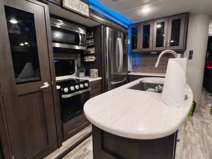 Well-equipped RV rental kitchen featuring dark cabinets, stainless steel appliances, an island sink, and blue accent lighting.. Grand Design Reflection 2020