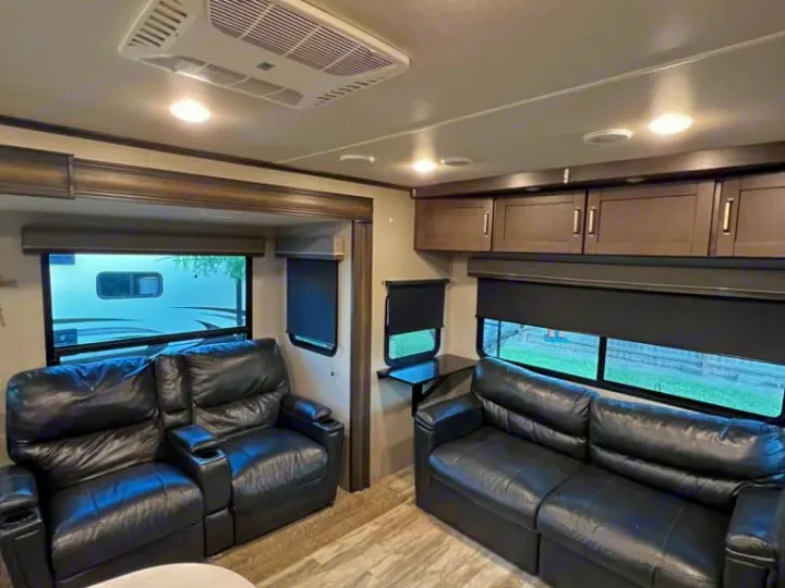 This spacious RV rental interior features dark leather recliners and a sofa, overhead cabinets, and large windows with blinds.. Grand Design Reflection 2020