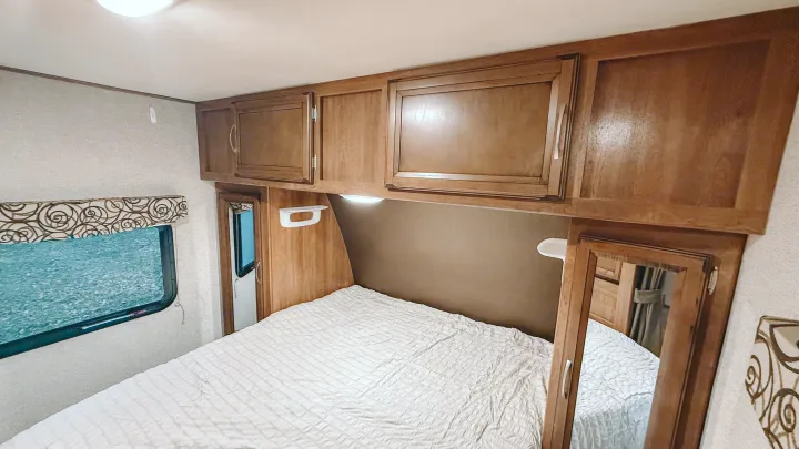 RV rental bedroom interior: light bedding, wood cabinets, small window, and a wall-mounted mirror. Cozy and compact space.. Coachmen Apex 2016