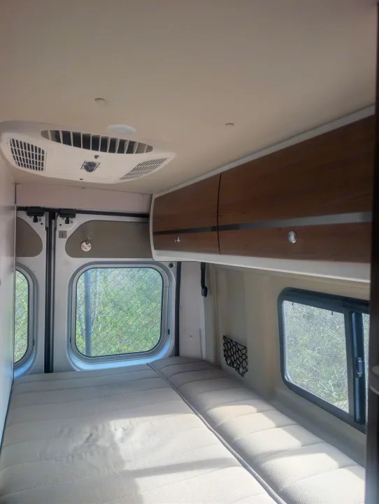 Cozy interior of an RV rental featuring a comfortable bed, windows, overhead cabinets, and an AC unit for your travels.. Winnebago Travato 2018