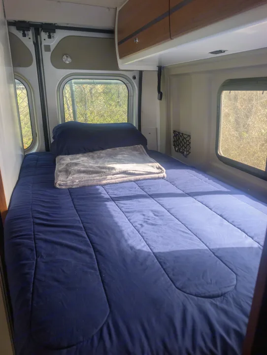 Comfortable bed setup in an RV rental, featuring blue bedding, a grey blanket, and sunlit windows for peaceful travels.. Winnebago Travato 2018