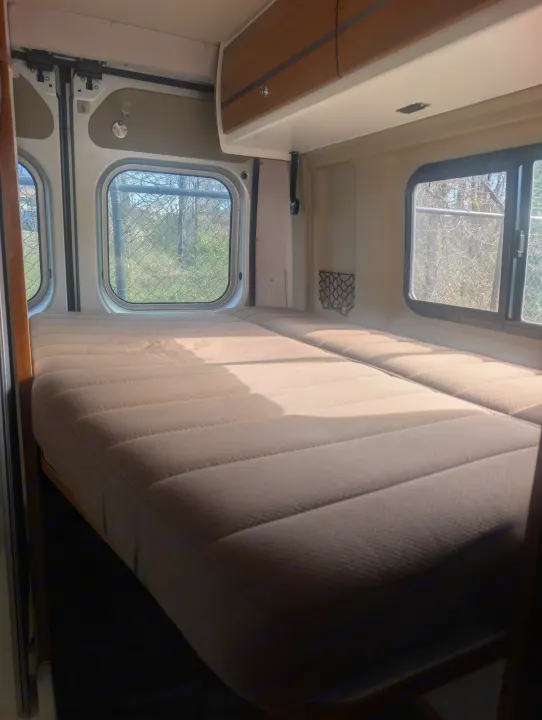 Bright rv rental interior with two beds, overhead cabinet, and side windows revealing trees and a fence outside, bathed in sunlight.. Winnebago Travato 2018