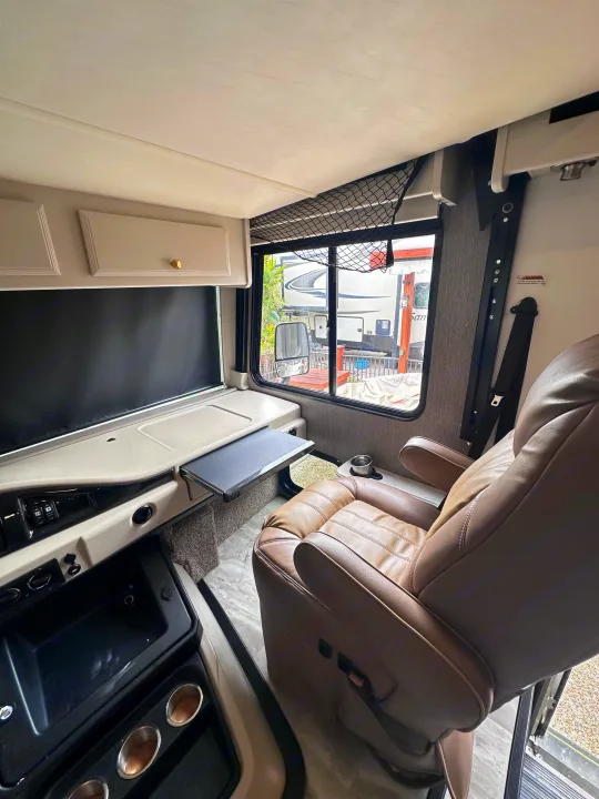 Spacious rv rental interior with a swiveled captain's chair, pull-out table, dashboard, and window view of other RVs.. Fleetwood Flair 2021