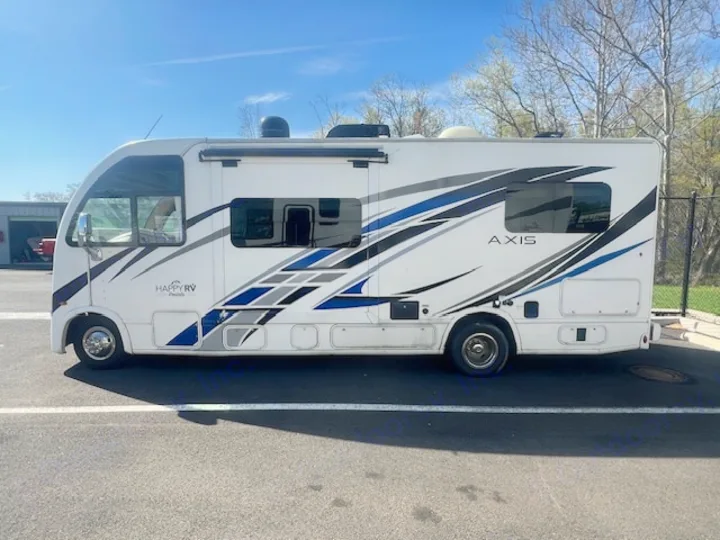 A white Thor Axis motorhome from Happy RV Rentals, featuring blue and grey graphics, parked on asphalt under a clear sky.. Axis 24.1 2027