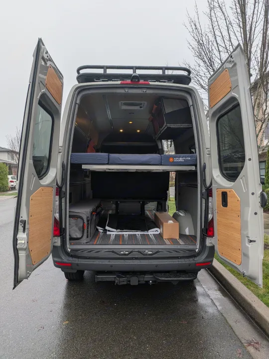 This custom camper van conversion is ideal for RV rental adventures, featuring an open rear with bed, storage, and roof rack.. Mercedes-Benz Sprinter 2025