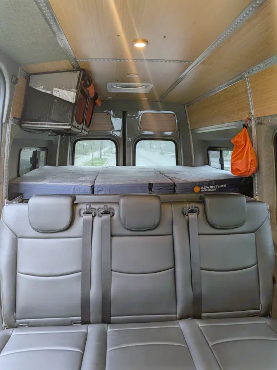 This rv rental interior features a 3-person black leather seat, custom bed with "Adventure Wagon" logo, wood-paneled ceiling, & windows showing a rainy day.. Mercedes-Benz Sprinter 2025