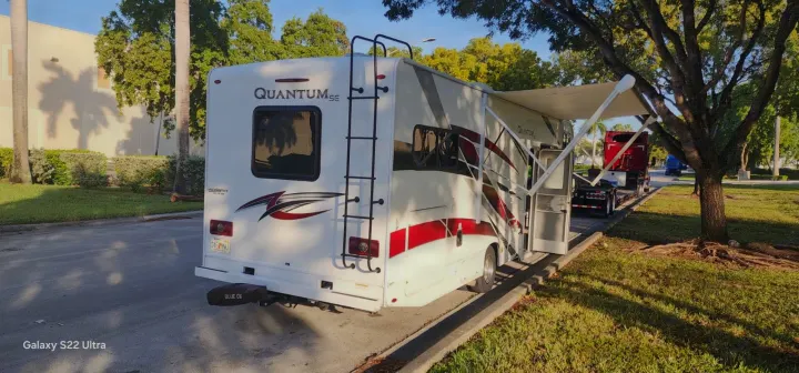 White Quantum SE RV rental with extended awning, parked outdoors. Rear view shows a ladder and a nearby semi-truck.. Thor Quantum 2021
