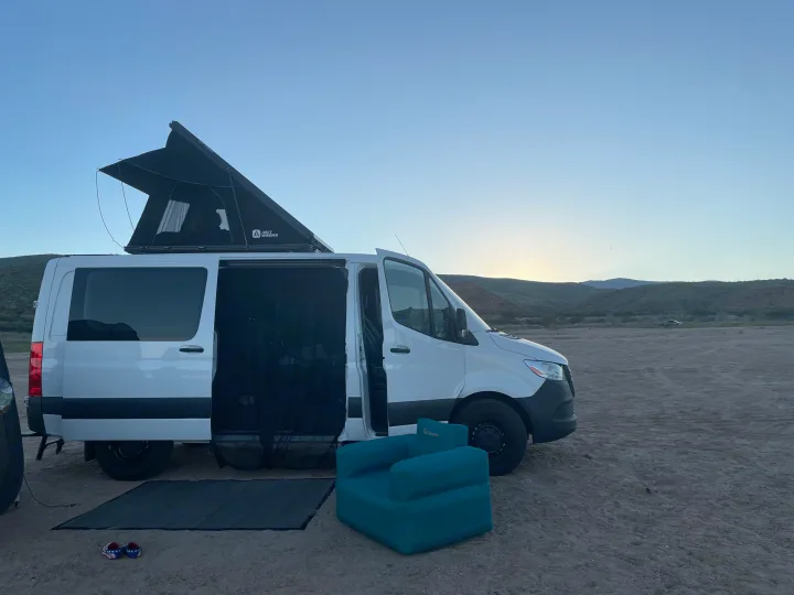 White rv rental camper van with pop-top tent, open door with mesh, teal chair, and mat on dirt. Desert sunset scene.. Mercedes Sprinter 2024