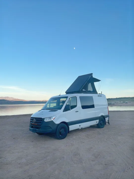 White RV rental van with a rooftop tent on a sandy lakeside at dusk, a crescent moon visible in the blue sky above mountains.. Mercedes Sprinter 2024