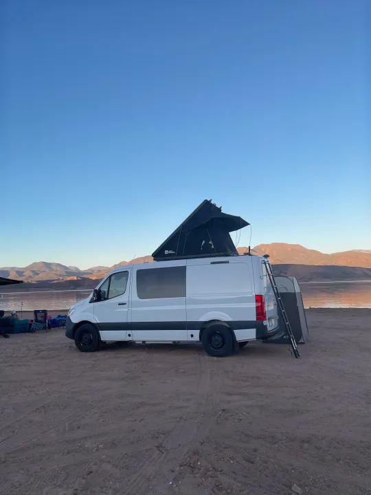 Enjoy nature with this white RV rental camper van, pop-top tent open, parked lakeside on sand, mountains under clear sky.. Mercedes Sprinter 2024
