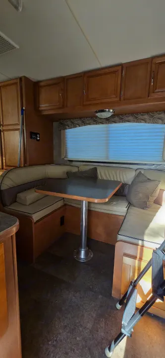 Light-toned U-shaped dinette with a dark table, overhead wood cabinets, and blinds in an RV rental interior.. Winnebago Minnie Winnie 2019