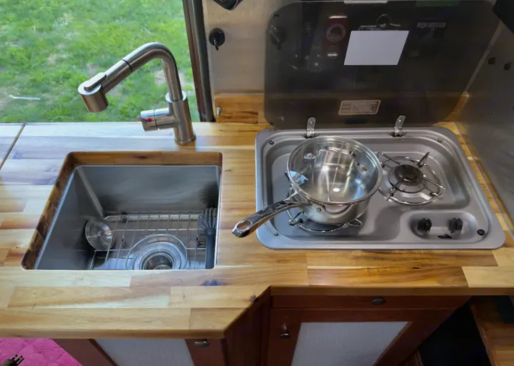 Compact RV rental kitchen: wood counter, stainless sink, modern faucet, and two-burner stove with a pot.. Roadtrek Simplicity SRT 2019