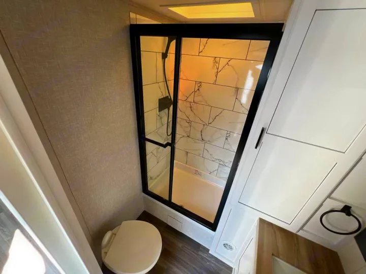 Clean RV rental bathroom: walk-in shower with black frame & marble-look tiles, toilet, white storage, and wood-look floor.. Jayco Eagle 2024
