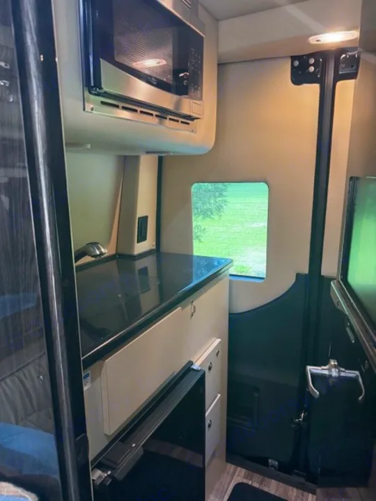 Inside an RV rental: Compact kitchen galley with microwave, black counter, and fridge drawers. Green view outside.. Ultimate Toys Ultimate Coach 2024