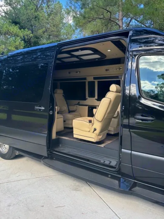 Explore this luxury RV rental's spacious interior! The black van has its side door open, revealing cream, plush captain's chairs.. Ultimate Toys Ultimate Coach 2024