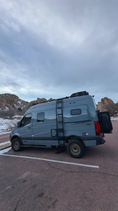 This gray Winnebago Revel adventure van, ideal for RV rental, is parked amidst snowy mountains under a cloudy sky.. Winnebago Revel 2023