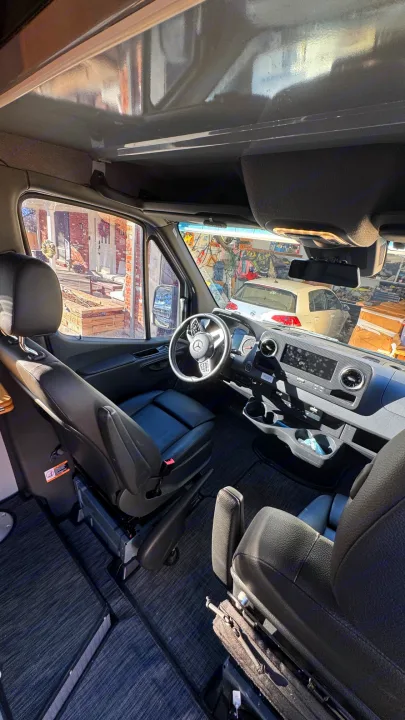 Inside this modern RV rental: A Mercedes Sprinter van's cockpit with sleek dashboard, black leather seats, and overhead storage.. Winnebago Revel 2023