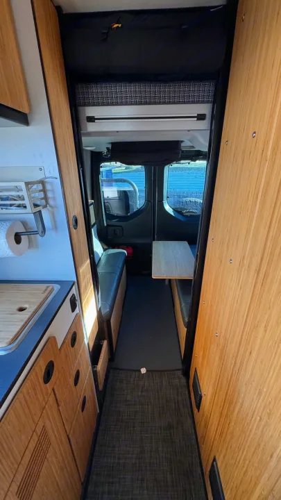 Interior view of an **RV rental** camper van, showcasing the kitchen counter, narrow aisle, and a cozy dining/seating area.. Winnebago Revel 2023