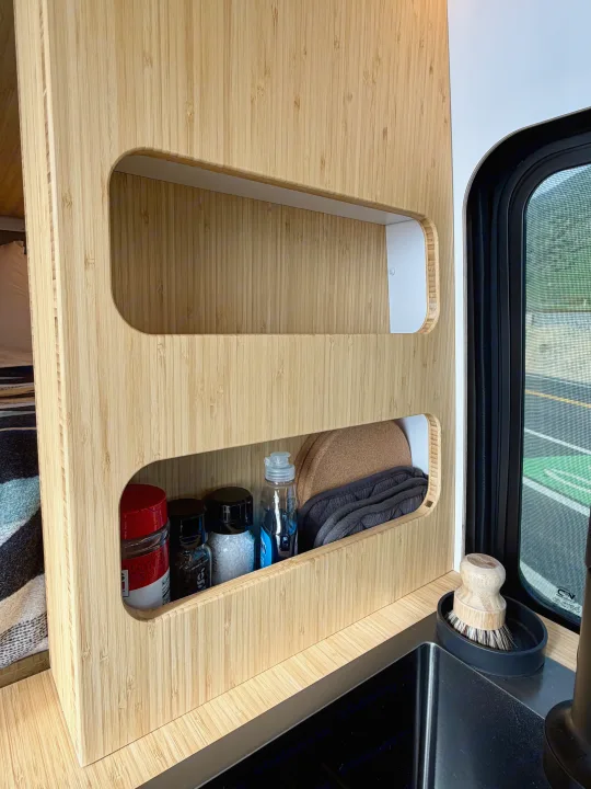 RV rental: Organized bamboo shelves in a van kitchen, holding spices, soap, and supplies by a sink and window.. Mercedes-Benz Sprinter 2025