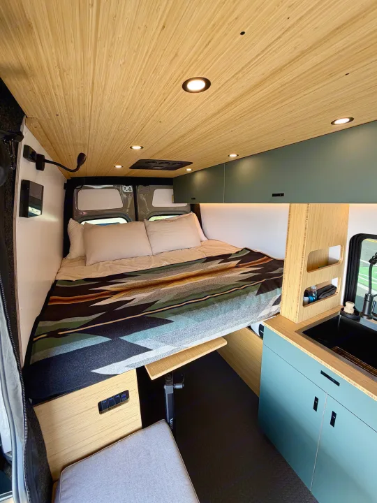 RV rental's modern interior: bamboo ceiling, cozy bed with patterned blanket, kitchenette & smart storage.. Mercedes-Benz Sprinter 2025