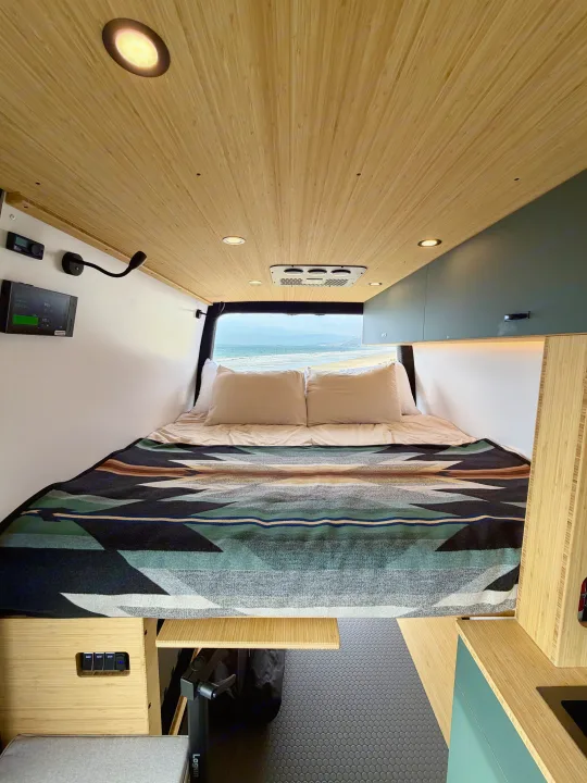 This rv rental shows a cozy camper van bed with a scenic ocean view, wooden ceiling, and a vibrant patterned blanket.. Mercedes-Benz Sprinter 2025