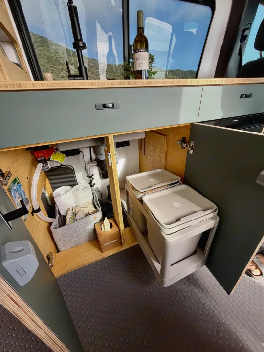 Organized RV rental kitchen interior featuring under-sink storage, pull-out bins for waste, & a scenic mountain view.. Mercedes-Benz Sprinter 2025