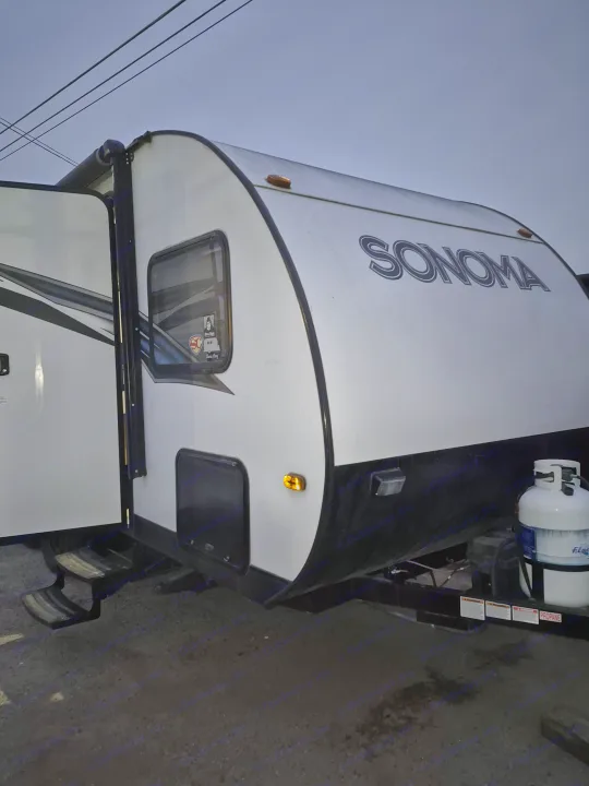 White Sonoma RV rental with an open door & steps, showing the front propane tank against a dim sky. Ideal for a travel adventure.. Forest River Sonoma 2020