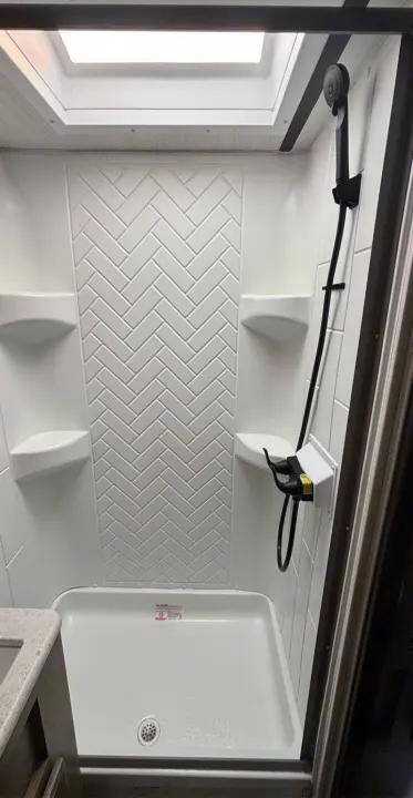 A clean, modern shower in an RV rental, with a skylight, white herringbone pattern wall, corner shelves, and black handheld showerhead.. Forest River Surveyor 2022