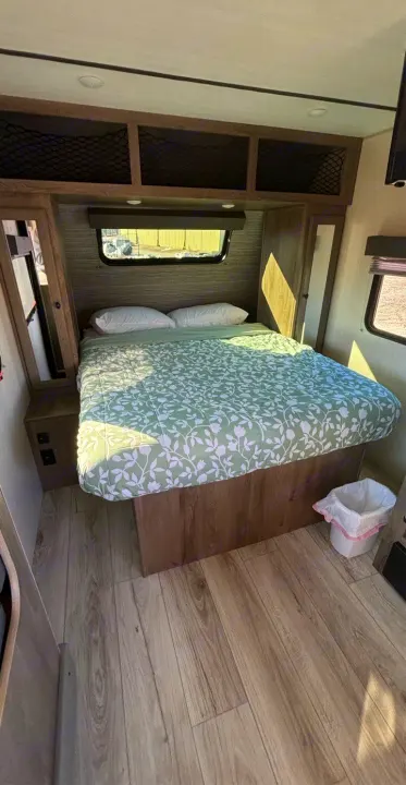 Cozy rv rental bedroom: green floral bed, overhead storage, window, and wood floor. Bright and inviting for your travels.. Forest River Surveyor 2022