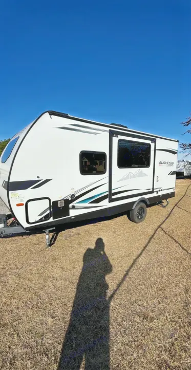 This white Surveyor Legend travel trailer, ready for rv rental, sits on dry grass under a clear blue sky. Person's shadow in foreground.. Forest River Surveyor 2022