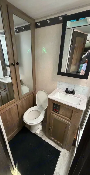 Compact RV rental bathroom with a white toilet, sink, and mirror. Wood-look cabinets and a black floor mat.. Forest River Surveyor 2022