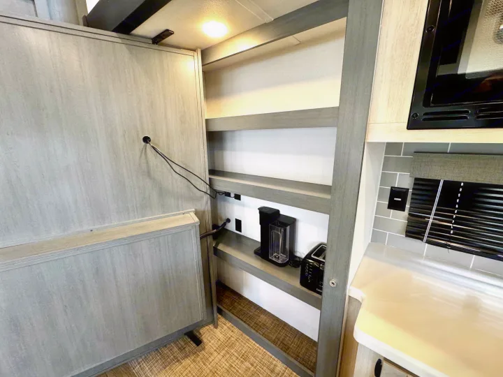 RV rental kitchen area: compact interior with microwave, shelves, coffee maker, toaster, countertop, and a folding wall on the left.. Forest River Puma Destination 2026