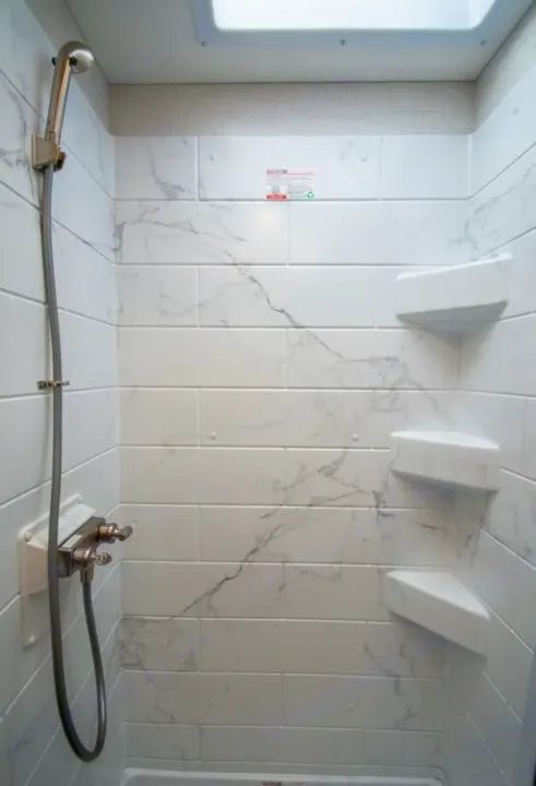 Clean, modern shower in an RV rental, featuring white marble-look tiles, a handheld showerhead, and corner shelves.. thor four winds 2027