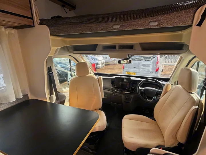 Bright RV rental interior: driver's cab with cream swiveling seats, black dining table, and an overhead bunk.. FORD TRANSIT RIMOR KILIG 669 5 BERTH 2026