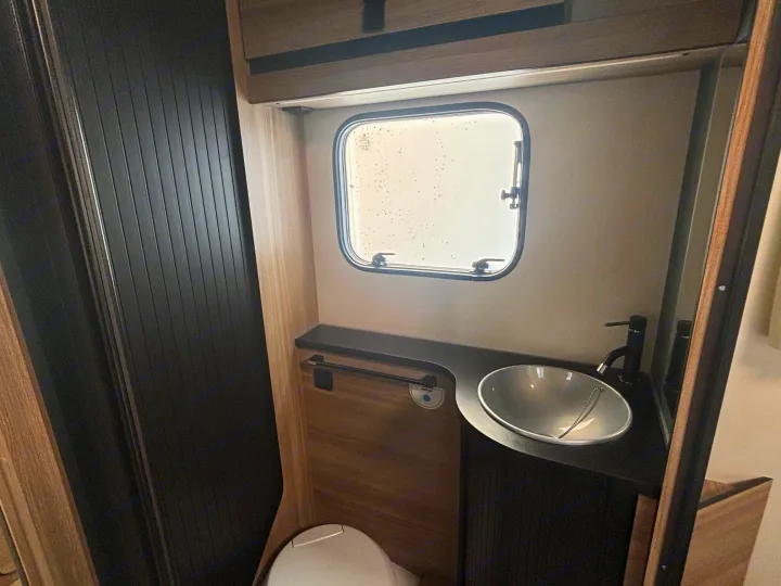 Compact RV rental bathroom features a toilet, stylish sink, modern faucet, and a small window in a dark wood interior.. FORD TRANSIT RIMOR KILIG 669 5 BERTH 2026