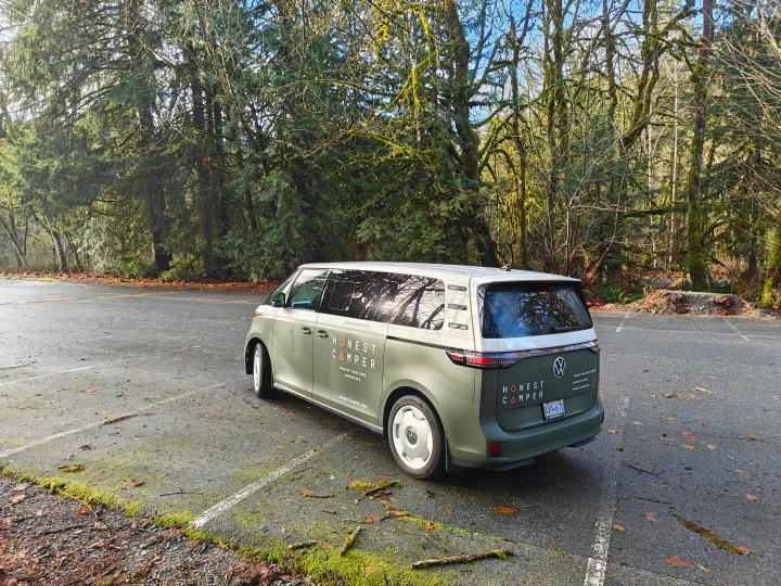 A green and white Honest Camper VW ID. Buzz rv rental van is parked in a mossy forest lot, ready for adventure.. Volkswagen ID. Buzz 2025
