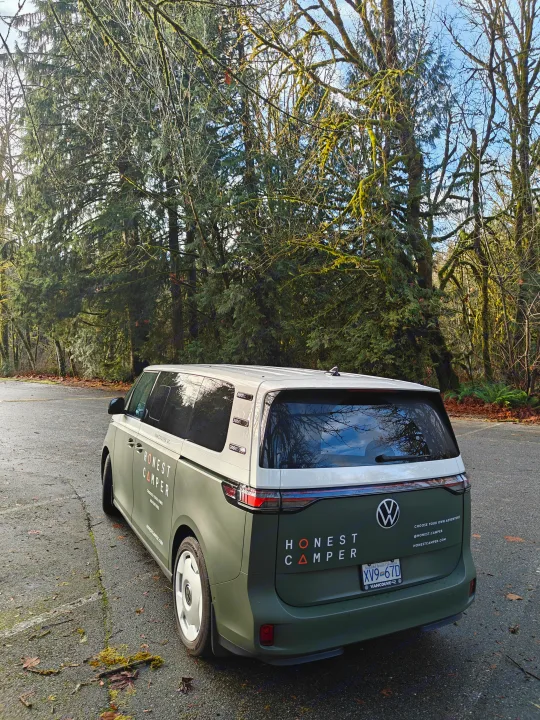 Green and white Honest Camper RV rental van parked beside lush, moss-covered trees in Vancouver, BC.. Volkswagen ID. Buzz 2025