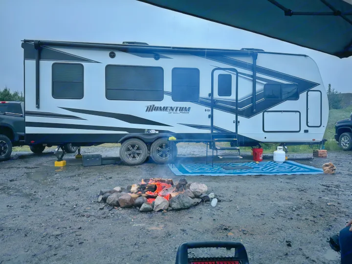 An RV rental, a Momentum G-Class travel trailer, is parked for camping, with a lit campfire nearby on wet ground.. Grand Design Momentum Toy Hauler 2022