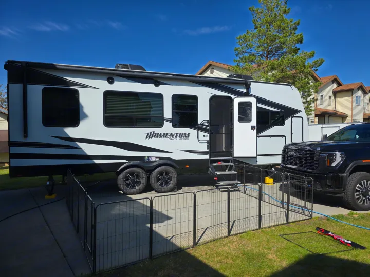 White Momentum G-Class RV rental (travel trailer) with a GMC truck and fenced area, parked on a sunny day.. Grand Design Momentum Toy Hauler 2022