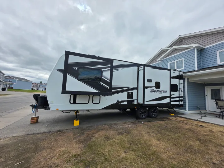 Modern white Momentum G-Class rv rental, with black accents, parked beside a blue house under a cloudy sky.. Grand Design Momentum Toy Hauler 2022